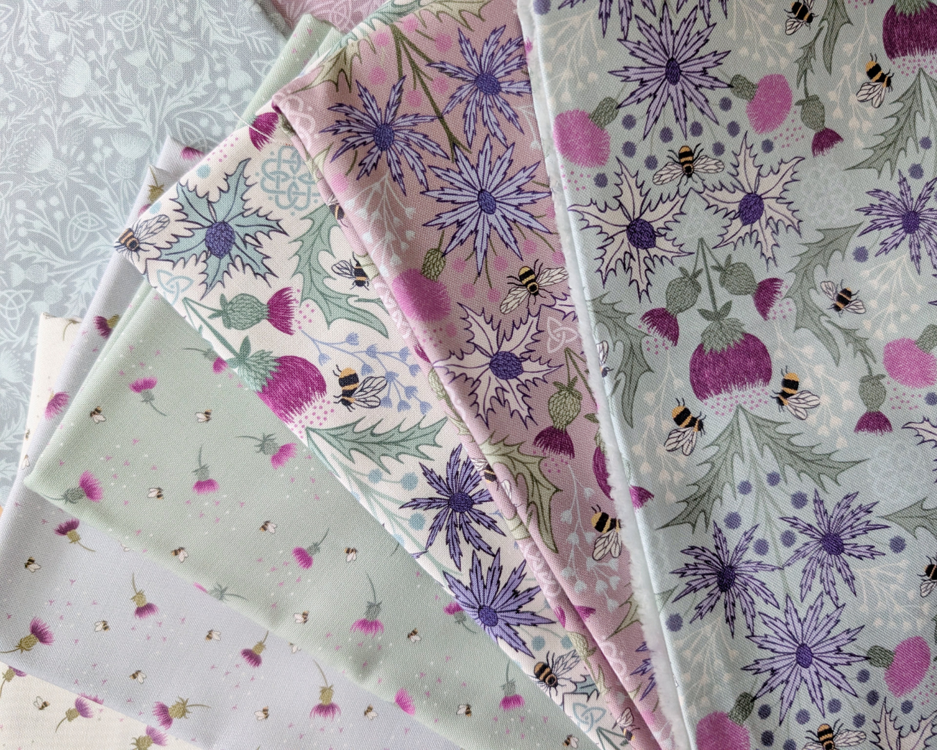 Stack of fabric swatches featuring Mirrored Thistle and Bee fabrics in cream, heather, and duck egg blue as the top three fabrics from left to right.