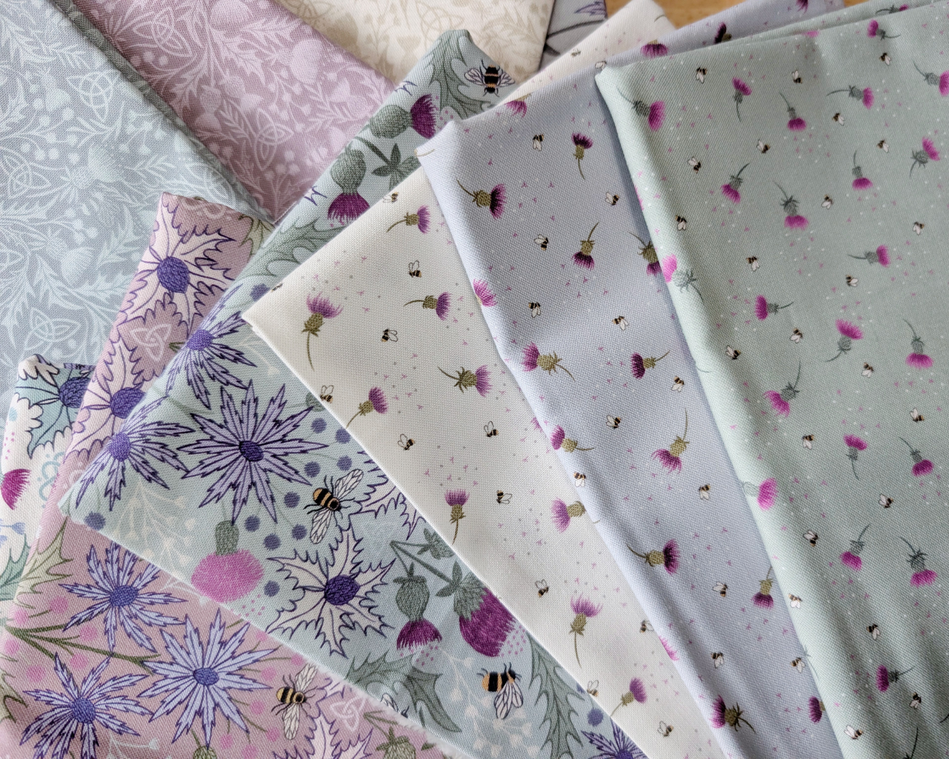 A stack of fabric swatches from A Celtic Summer collection, displaying Bee Thistle in cream, sky blue, and sage green from left to right as the top three fabrics.